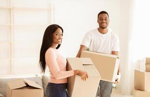 Moving into new home concept. Happy black guy and his beautiful girlfriend with boxes posing and smiling during relocation. Young African American couple unpacking things together photo