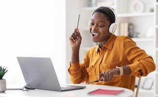Joyful short-haired millennial black lady having break while working at office, watching on laptop, using wireless headset, holding pens and playing imaginary drums, relaxing, copy space photo