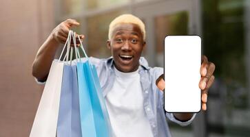 Unbelievable bonus for mobile user, online shopping application. Surprised african american man with packages shows smartphone with blank screen to camera photo