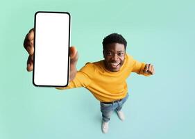 Excited black guy demonstrating smartphone with blank white screen, showing free copy space for your ad, recommending mobile app or website, standing over blue background, mockup photo