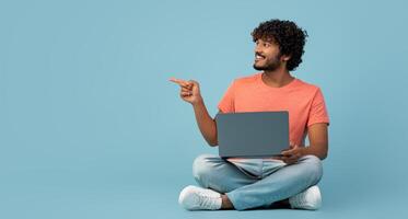 Handsome millennial indian man in casual outfit sitting on floor, using modern laptop on blue studio background, typing on computer keyboard, working online, pointing at copy space, panorama photo