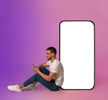 Cheery millennial Arab guy sitting near big cellphone with blank white screen, using mobile device, using new app in neon light, mockup for website or application design photo