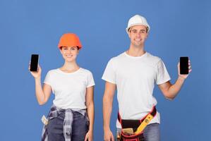 Positive couple of repair workers in protective hardhats holding smartphones with black screen, demonstrating empty copy space for website or app design, standing over blue studio background, mockup photo