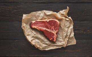 Raw t-bone steak on craft paper on dark background. Prime meat on black table with spices, copy space, top view photo