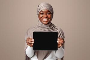 Mockup Image Of Smiling African Muslim Lady Holding Digital Tablet With Black Screen, Islamic Woman In Hijab Demonstrating Device With Copy Space For Your Website Or App Design Over Light Background photo
