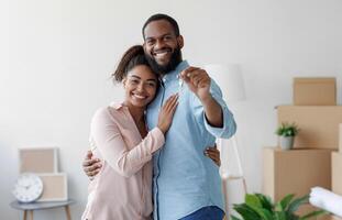 Happy smiling young african american loving family hugging in new home among boxes and showing house keys, man and woman spouses feel joy. Moving, relocating to own apartment, rental and real estate photo