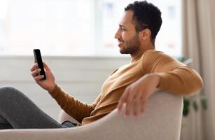 Profile side view portrait of young smiling Arab man holding mobile phone sitting on the couch in living room. Cool guy browsing internet, surfing web or watching , using application, free space photo