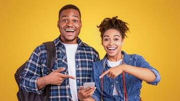 Excited Black Travelers Couple Pointing Finger At Smartphone Recommending Mobile Travel Application Standing Over Yellow Background In Studio. Approving App For Tourists Concept photo