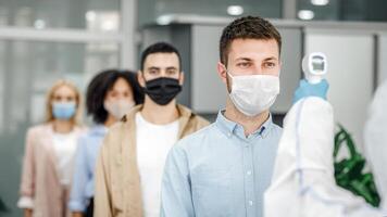 Morning at work during covid-19 epidemic. Person with contactless thermometer measures temperature of multiracial employees in protective masks an entrance of modern office, copy space photo