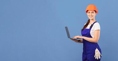 Cheerful young lady in coveralls and hard hat using laptop computer on blue background in studio, smiling female worker ordering building instruments and materials for repairment online, copy space photo