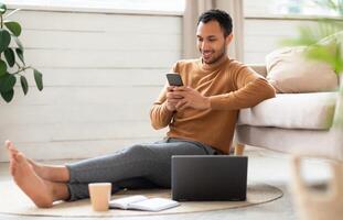 Portrait of young man holding using cell phone sitting on floor leaning on couch in living room. Cool guy browsing internet, surfing web watching , using application and pc, full body length photo