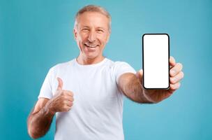 I Recommend. Portrait of smiling mature man holding smartphone with white blank screen in hand showing device and thumb up gesture. Selective focus on gadget with empty free space for mock up template photo
