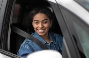 Great auto offer, car advertisement and modern blogger. Smiling happy young african american woman sitting in transport behind steering wheel with seat belt and looking out open window, empty space photo