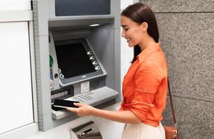 Young lady customer using smartphone to gain access to ATM, withdrawing money from account standing near bank machine on a street outside, side view shot. Financial safety and transactions photo