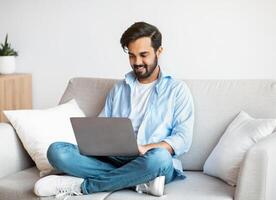Distant Work. Happy Arab Freelancer Man Using Laptop Computer At Home, Typing On Keyboard, Young Eastern Guy Sitting On Sofa In Living Room, Working Remotely On Online Project, Copy Space photo