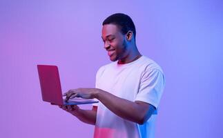 Modern Technologies. Happy Black Man Using Laptop Computer In Neon Light, Enjoying Online Communication, Gaming And Remote Job Offers, Excited Guy Standing Over Purple Studio Background, Copy Space photo