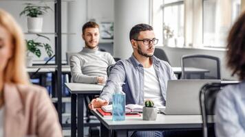 Creating ideas to work after quarantine. Young attractive man in glasses looks out the window, sitting at table with laptop, plant in pot and antiseptic with colleagues in interior of office, empty space photo