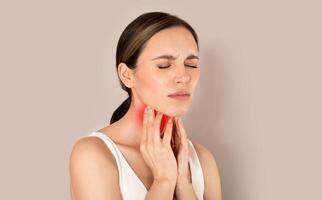 Unhappy young brunette woman with closed eyes wearing white top suffering from pain in throat, touching red inflamed zone on her neck, panorama with empty space, beige studio background photo
