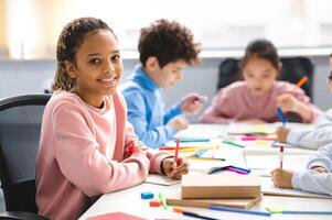 Academic Concept. Smiling junior african american school girl sitting at desk in classroom, writing in notebook, posing and looking at camera. Group of classmates studying in the background photo