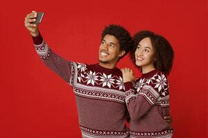 Guy and lady in identical New Year outfits happily pose for selfie. Smiling young african american couple watching in smartphone, make photo for Christmas card, isolated on red background, studio shot