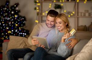 Delighted husband and wife making purchases online, using digital tablet and credit card, Christmas atmosphere at home. Happy young family shopping online on pad, using Christmas offers, copy space photo