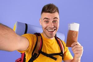 Happy young guy with backpack, passport and plane tickets taking selfie on blue background photo