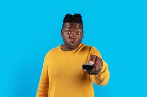 Shocked Black Man Watching Television, Pointing Remote Controller At Camera, Amazed Young African American Guy Switching Tv Channels While Standing Isolated Over Blue Background, Copy Space photo