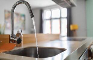 Close up shot of modern kitchen faucet with water running from tap, flow falling to empty dish sink at stylish home interior, selective focus, sustainable resources consumption concept photo