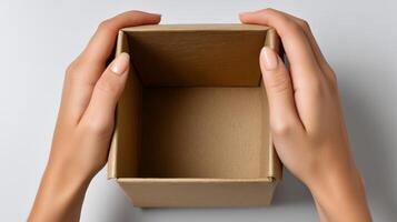 Two hands grasp an empty cardboard delivery box set on a smooth surface. The box is open, indicating the anticipation of unpacking new items. The setting is minimal and organized. photo
