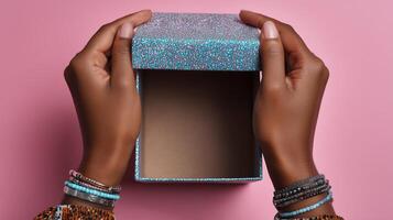 A pair of hands gently lifting the lid off a vibrant box featuring a textured surface. The box rests on a soft pink background, showcasing the anticipation of discovering its contents. photo