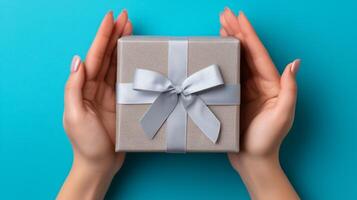 Two hands gently cradle an open delivery box adorned with a silver ribbon against a bright turquoise background. The box appears ready for gifting or reveal, suggesting excitement and anticipation. photo