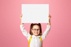 Preparation for school. Smiling little girl with backpack in glasses, holding sign with empty space, isolated on pink background, studio shoot photo
