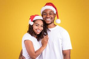Loving African Couple In Christmas Santa Hats Posing Hugging Over Yellow Background In Studio, Smiling To Camera. Xmas And New Year Joy, Winter Holidays Celebration Concept. photo