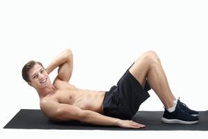 Smiling millennial guy with naked chest doing abs exercises on sports mat over light studio background, panorama. Full length portrait of athletic man working out core muscles, keeping in good shape photo
