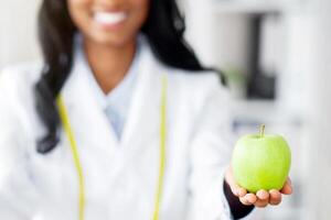 Closeup view of dietitian's hands holding fresh apple and donut at table in medical office. Cropped view of black nutritionist offering choice between healthy and junk foods. Dieting concept photo