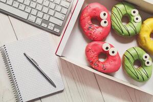 Box of colorful glazed donuts with funny eyes on the office desk near computer keyboard and notepad with pen. White rustic wooden table with business lunch snack dessert top view photo