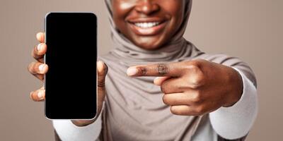 Unrecognizable African Muslim Lady Pointing Finger At Smartphone With Black Blank Screen In Her Hand, Demonstrating Empty Mockup Place For Your App Or Website Design, Cropped Image, Panorama photo