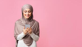 Millennial muslim woman holding smartphone in hands, texting, reading message, or using new application, standing over pink background with free space photo