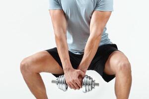 Strength workout concept. Positive young sportsman with dumbbell doing squat on light studio background, full length. Handsome bodybuilder lifting weights, doing powerlifting exercises photo