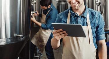 Beer production process at brewery. Man operating brewing equipment at beer making factory and using digital tablet. Cheerful young handsome men in aprons check data on large kettle with craft drink photo