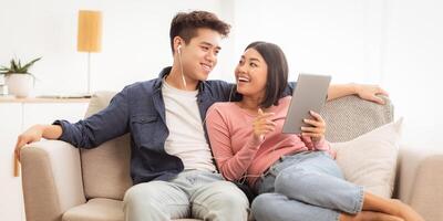 Japanese Spouses Using Digital Tablet Computer Browsing Internet Online Sitting On Couch In Living Room At Home. Girlfriend And Boyfriend Relaxing Spending Weekend Together. Modern Gadgets And Apps photo