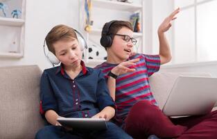 Boys in headphones enjoying music from laptop and tablet. Friends having fun at home photo