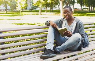 Smiling african-american student reading a book in the park, studying and preparing for exams at the university or college. Technology, communication, education and remote working concept, copy space photo