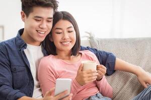 Korean Couple Using Smartphone Relaxing Sitting On Sofa At Home On Weekend. Cellphone Users Browsing Internet, Gadget Lifestyle, Mobile Application Concept. Empty Space photo
