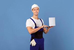 Smiling young man in coveralls and hard hat using laptop computer on blue studio background, handsome male worker ordering building instruments and materials for flat renovation online, copy space photo