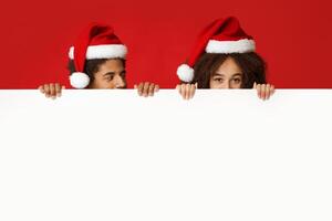 New Year banner and Merry Christmas. Mysterious young african american male and female in Santa hats, holds white billboard with empty space and looks out from behind it, isolated on red background photo