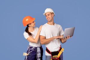 Cheerful young couple in protective hardhats using laptop computer while posing on blue studio background, checking renovation project for their apartment, searching repair tools online in internet photo