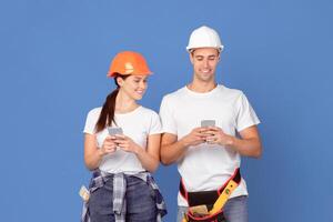 Smiling Man And Woman In Hard Hats Using Smartphones Over Blue Background, Browsing New App, Having Rest While Repair House Together Or Shopping Construction Tools Online, Copy Space photo
