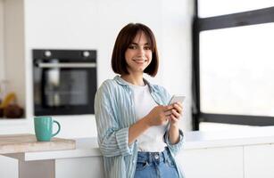 Smiling young lady using smartphone while enjoying coffee in kitchen interior at home, standing near table and smiling at camera, copy space photo