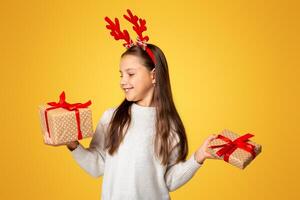 Dreams come true on Christmas. Smiling little kid girl with horns in sweater holds presents in festive package, isolated on yellow background photo